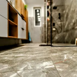 Szare płytki podłogowe imitujące marmur z pomarańczowymi żyłkami w nowoczesnej łazience z przeszklonym prysznicem i szafkami z drewnianymi akcentami.