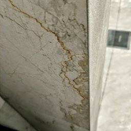 Dan72 - Fragment narożnika ściany wyłożonej płytkami imitującymi marmur z wyraźnymi, brązowymi żyłkami, widoczne fugi i fragment podłogi w tle.