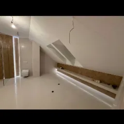 Wykończone poddasze ze skośnym sufitem, jasną podłogą i oświetloną półką wnękową, widoczna kabina prysznicowa i wisząca żarówka bez klosza.