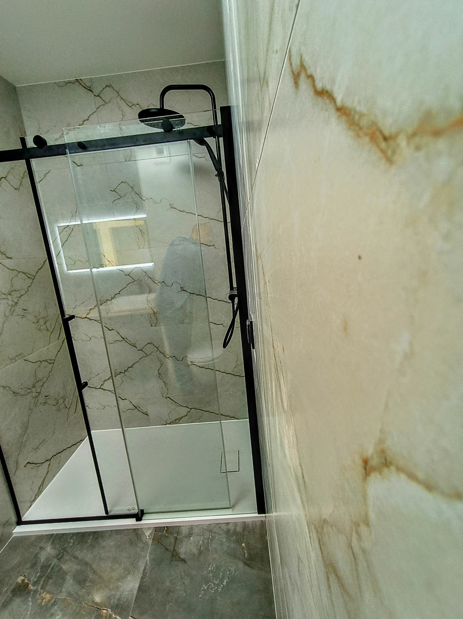 Kabina prysznicowa z czarnymi profilami i deszczownicą zamontowana w rogu łazienki, wykończonej płytkami imitującymi marmur w odcieniach szarości i beżu.