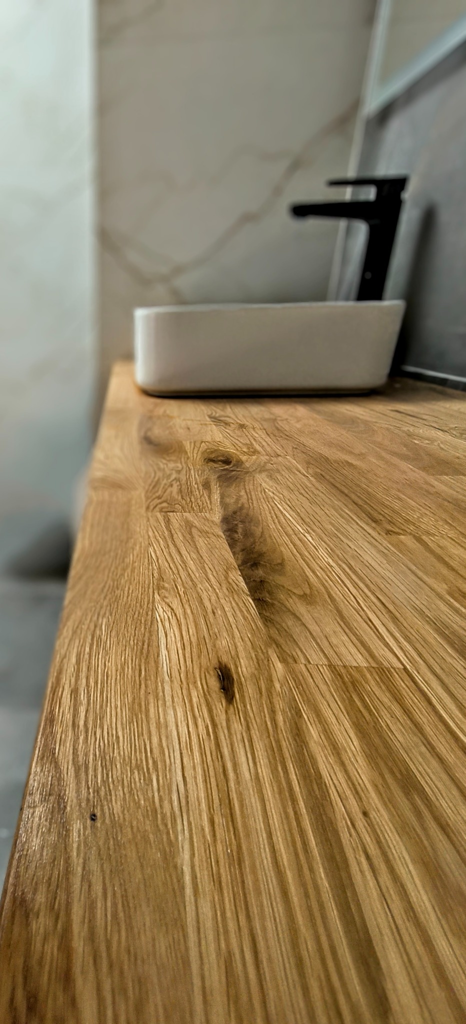 Blat dębowy pod umywalkę nablatową, widoczne usłojenie drewna i czarny kran w tle, marmurowe płytki na ścianie.