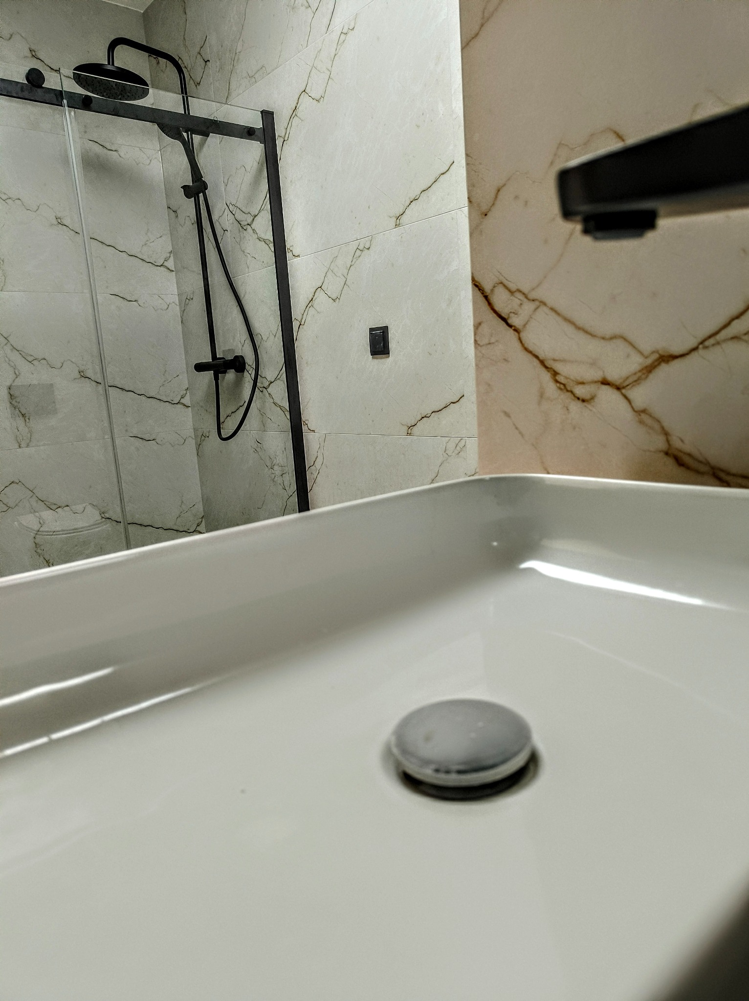 Nowoczesna łazienka z białą umywalką nablatową, czarną armaturą i kabiną prysznicową z marmurowymi płytkami na ścianach.