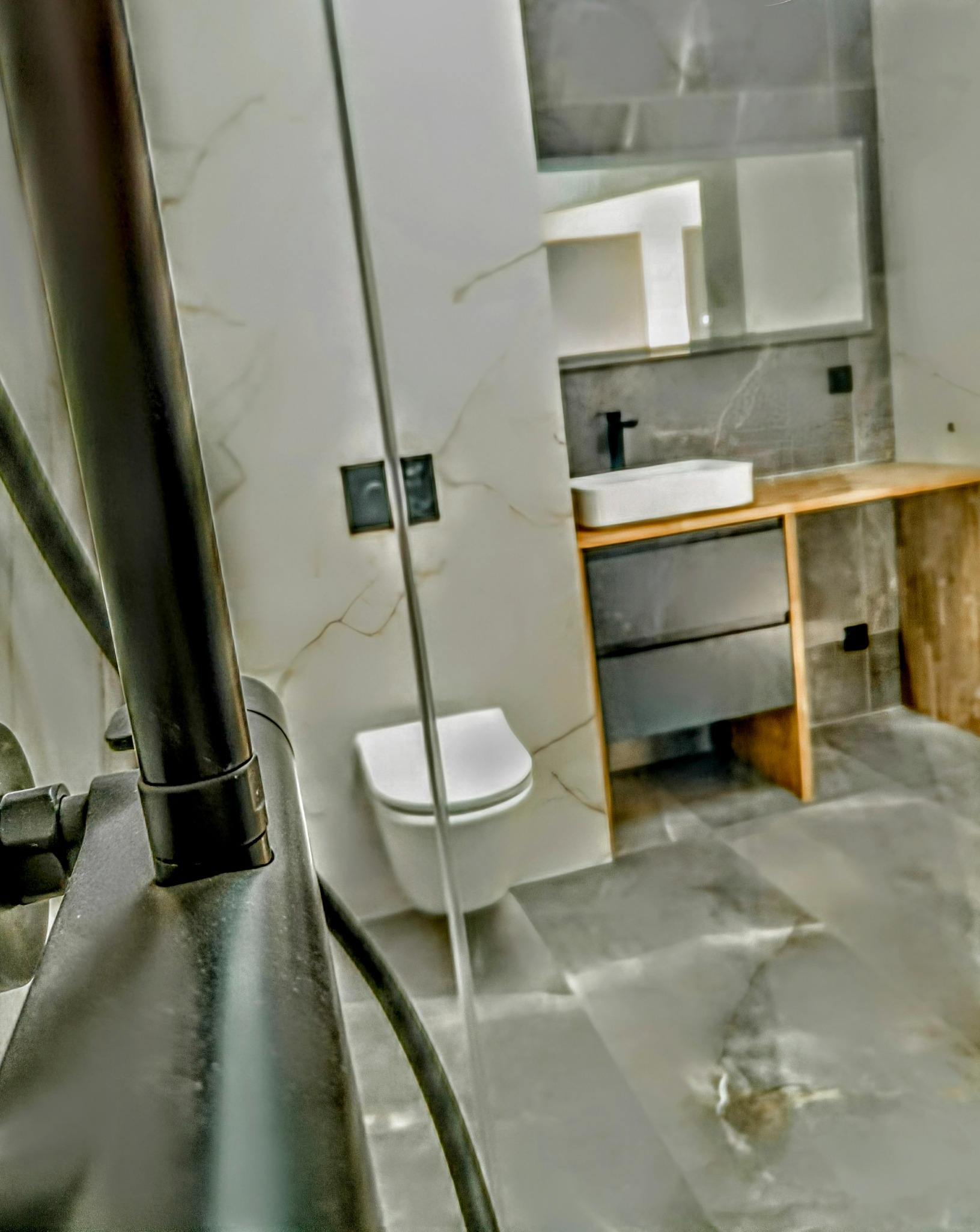 Nowoczesna łazienka z kabiną prysznicową typu walk-in, marmurowymi płytkami na ścianach i podłodze, wiszącą toaletą oraz umywalką nablatową z czarną armaturą.