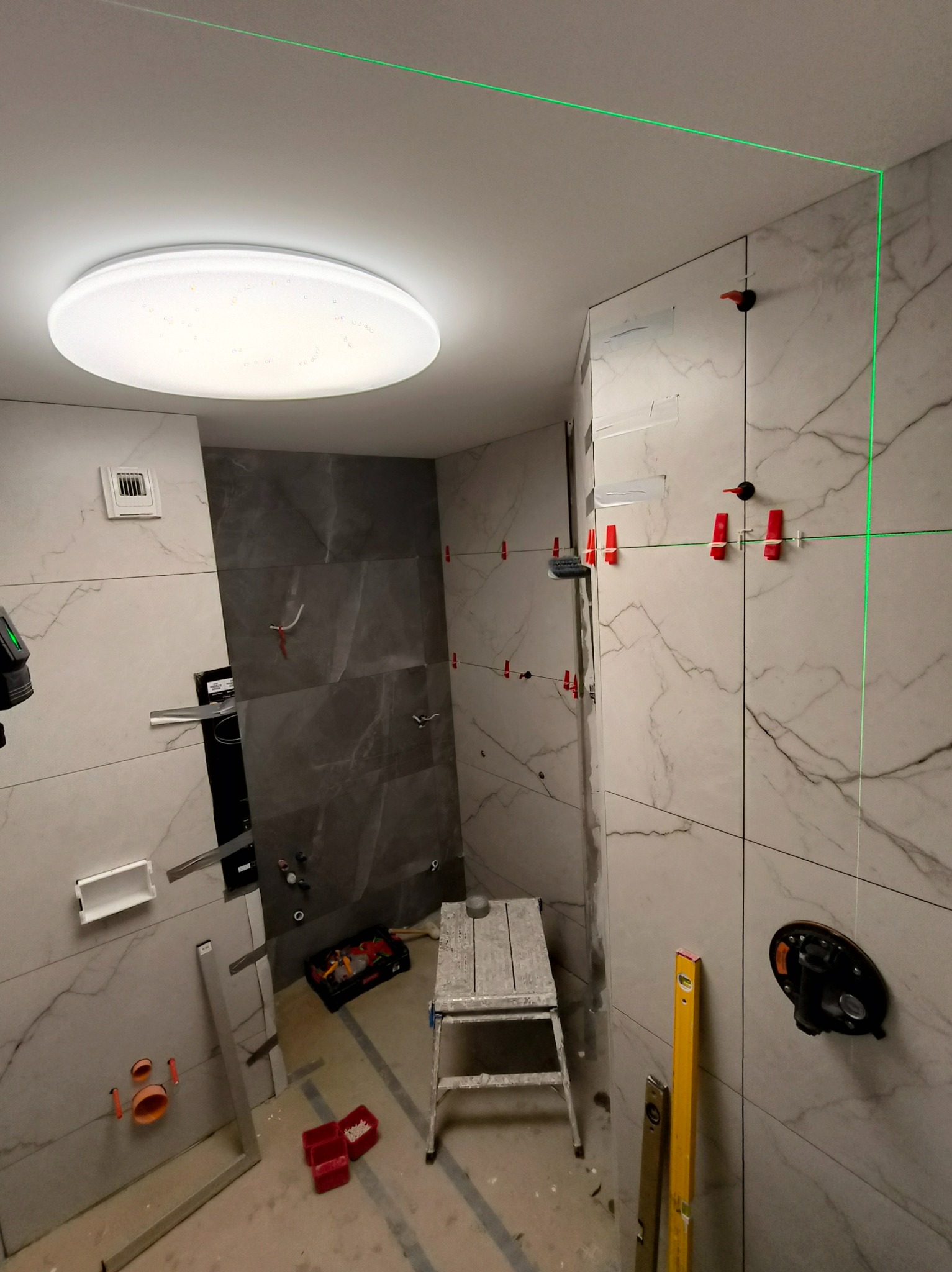 Łazienka w trakcie prac wykończeniowych; widoczne płytki imitujące marmur, poziomica laserowa wyznaczająca linię, krzyżyki dystansowe i narzędzia.