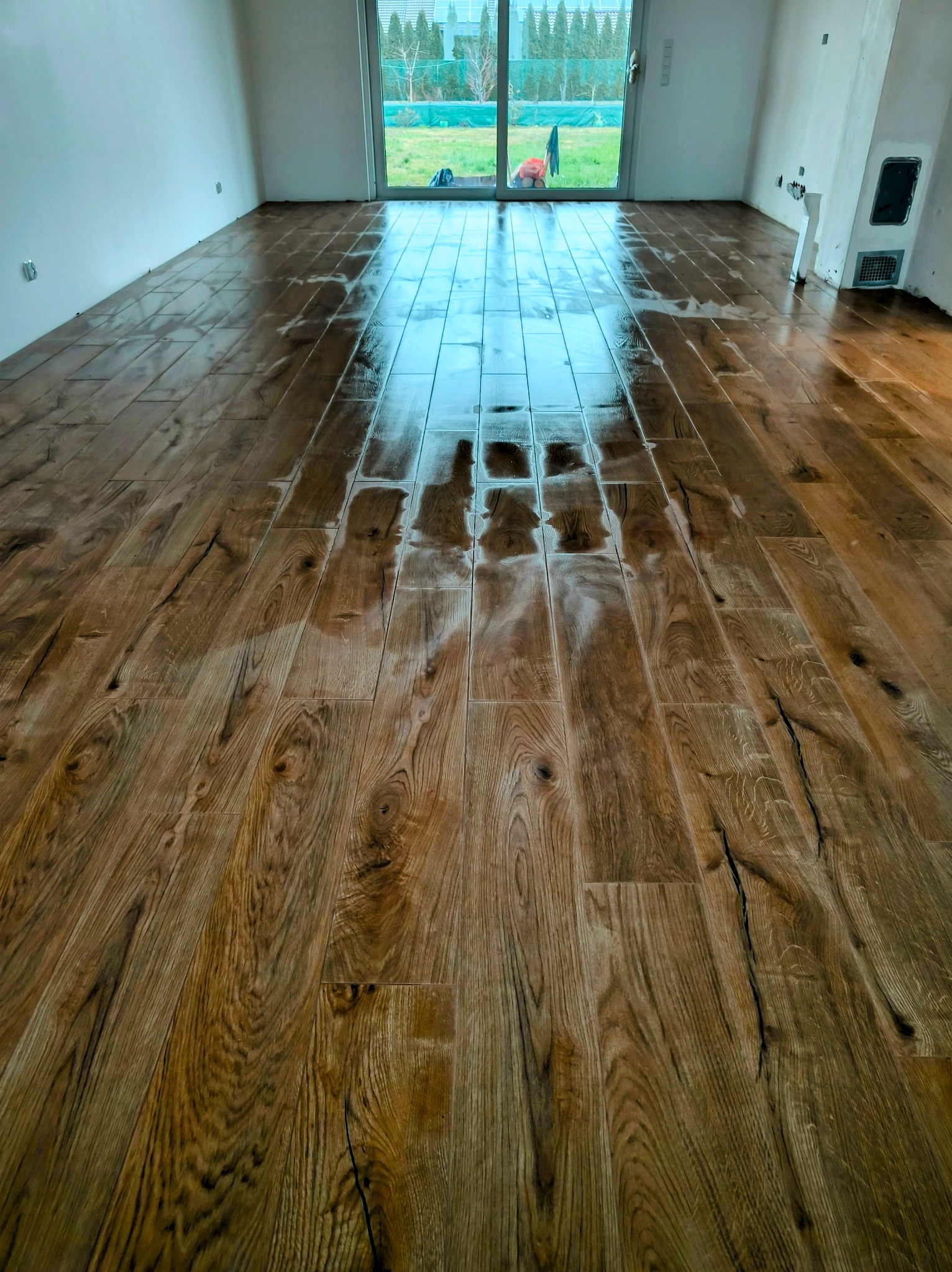 Świeżo polakierowana, błyszcząca drewniana podłoga ułożona w pustym pokoju z dużym oknem balkonowym i niedokończonymi ścianami.
