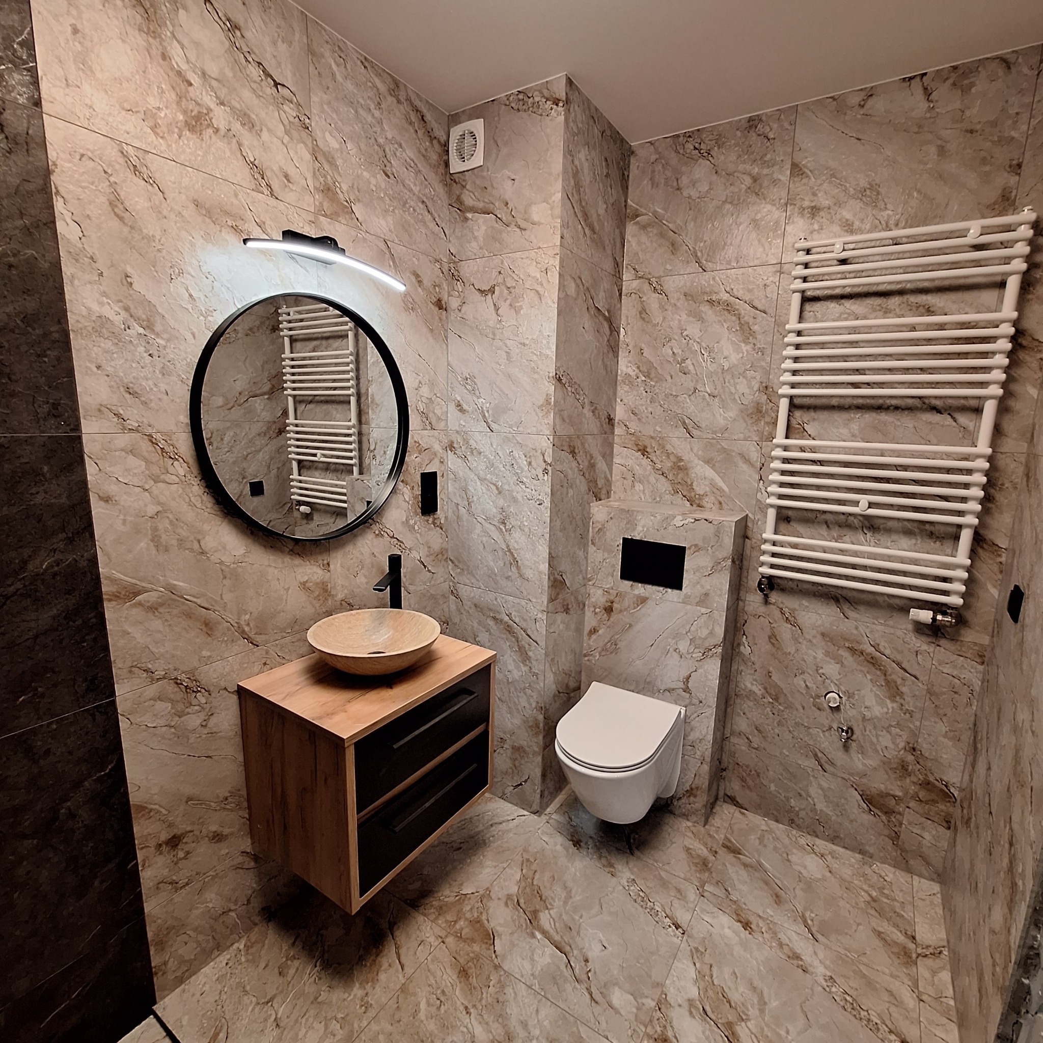 Nowoczesna łazienka z marmurowymi płytkami, okrągłym lustrem z podświetleniem LED, drewnianą szafką pod umywalką, wiszącą toaletą i grzejnikiem drabinkowym.