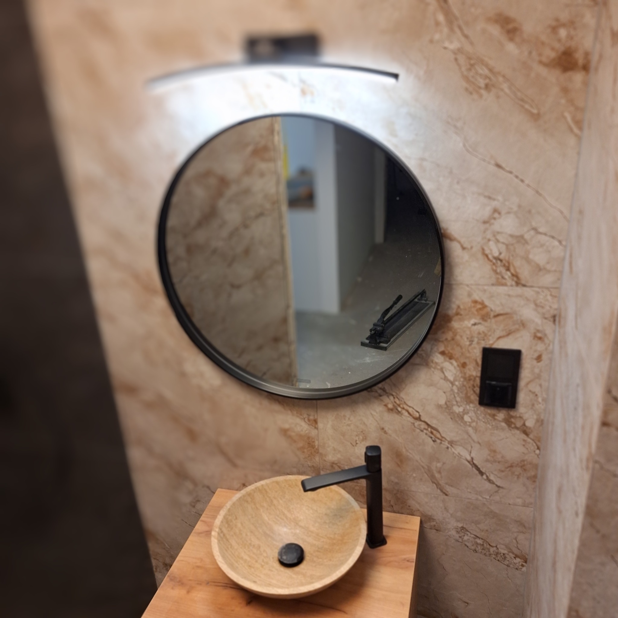 Nowoczesna łazienka z okrągłym lustrem z podświetleniem LED, drewnianą umywalką i czarną armaturą na tle ściany wyłożonej płytkami imitującymi marmur. W lustrze odbija się narzędzie do cięcia płytek.