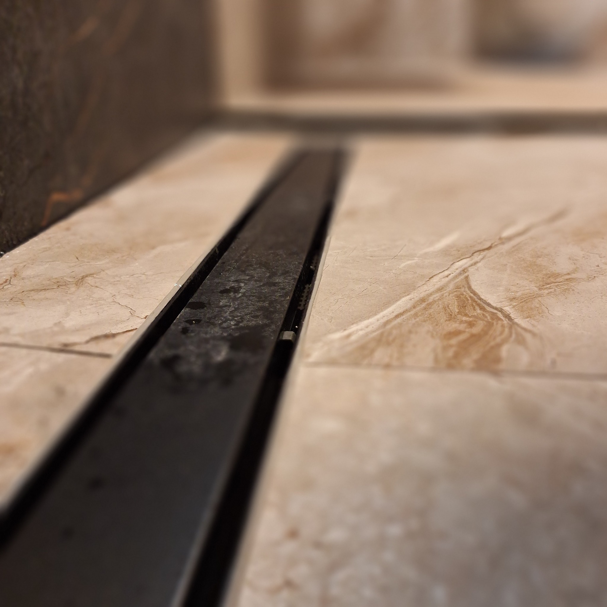 Szczegółowe ujęcie liniowego odpływu prysznicowego wkomponowanego w beżowe płytki podłogowe o marmurkowym wzorze, z widocznymi drobnymi zaciekami w odpływie.