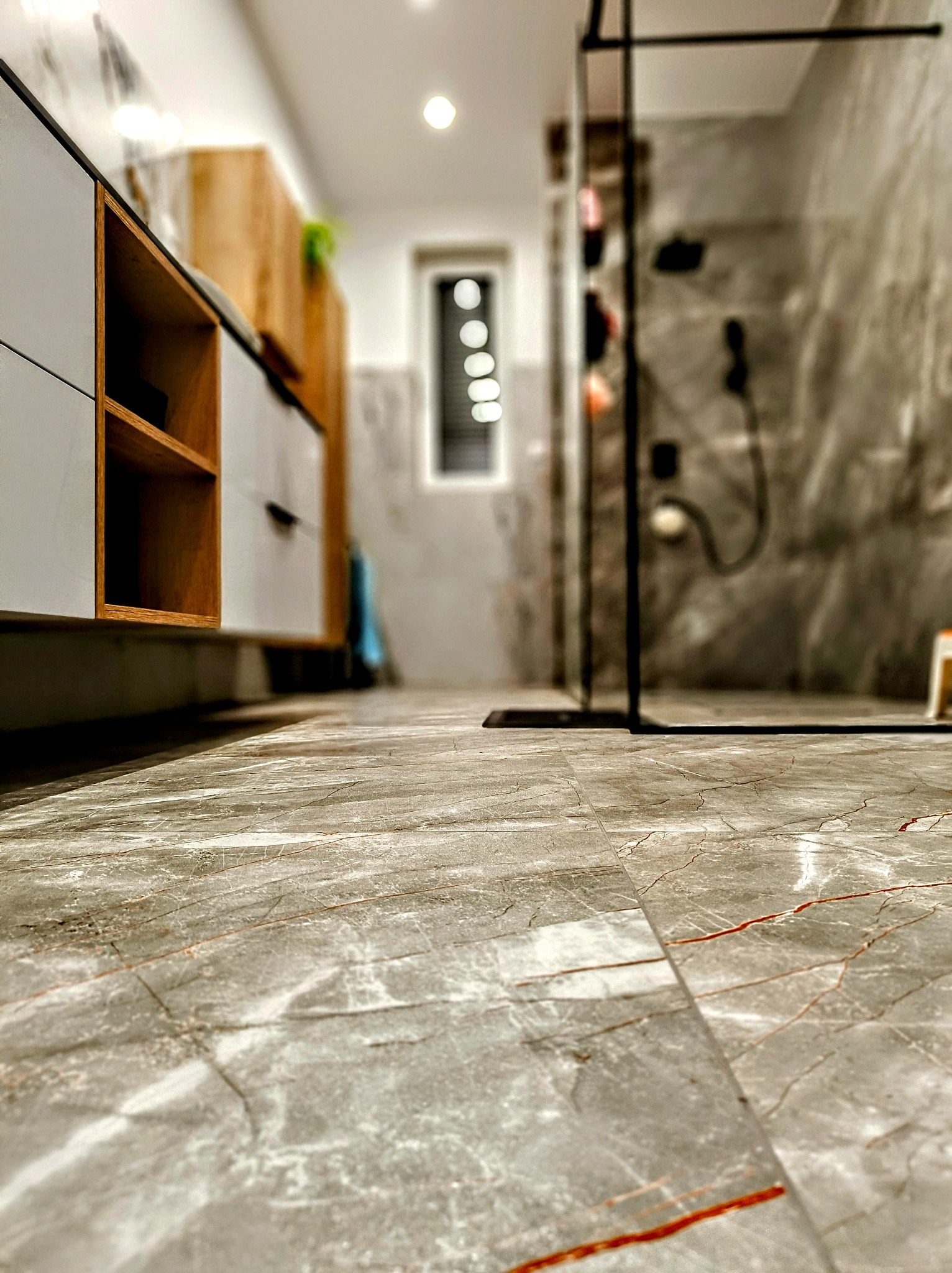 Szare płytki podłogowe imitujące marmur z pomarańczowymi żyłkami w nowoczesnej łazience z przeszklonym prysznicem i szafkami z drewnianymi akcentami.