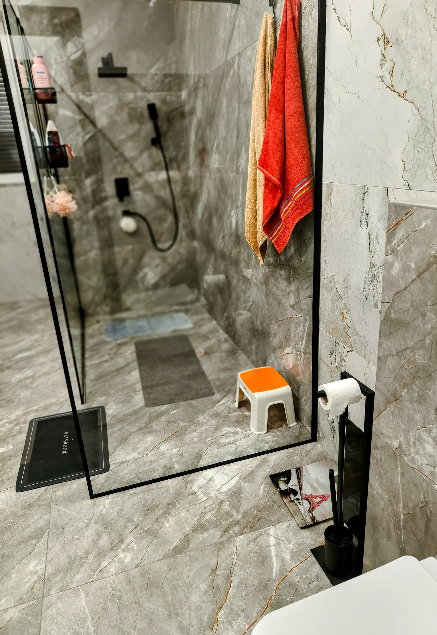 Nowoczesna łazienka z marmurowymi płytkami, kabiną prysznicową typu walk-in, ręcznikami w odcieniach beżu i czerwieni oraz pomarańczowym stołkiem.