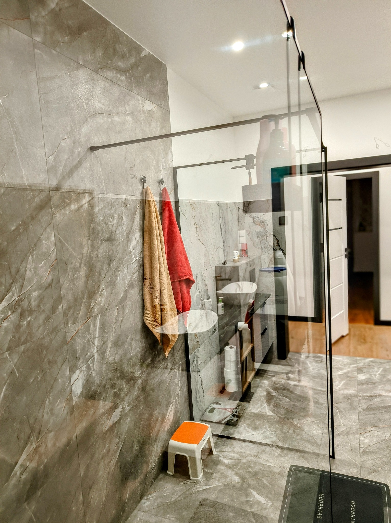 Nowoczesna łazienka z marmurowymi płytkami, szklaną kabiną prysznicową typu walk-in, dwoma umywalkami na blacie i ręcznikami na wieszakach, oświetlona punktowymi lampami sufitowymi.