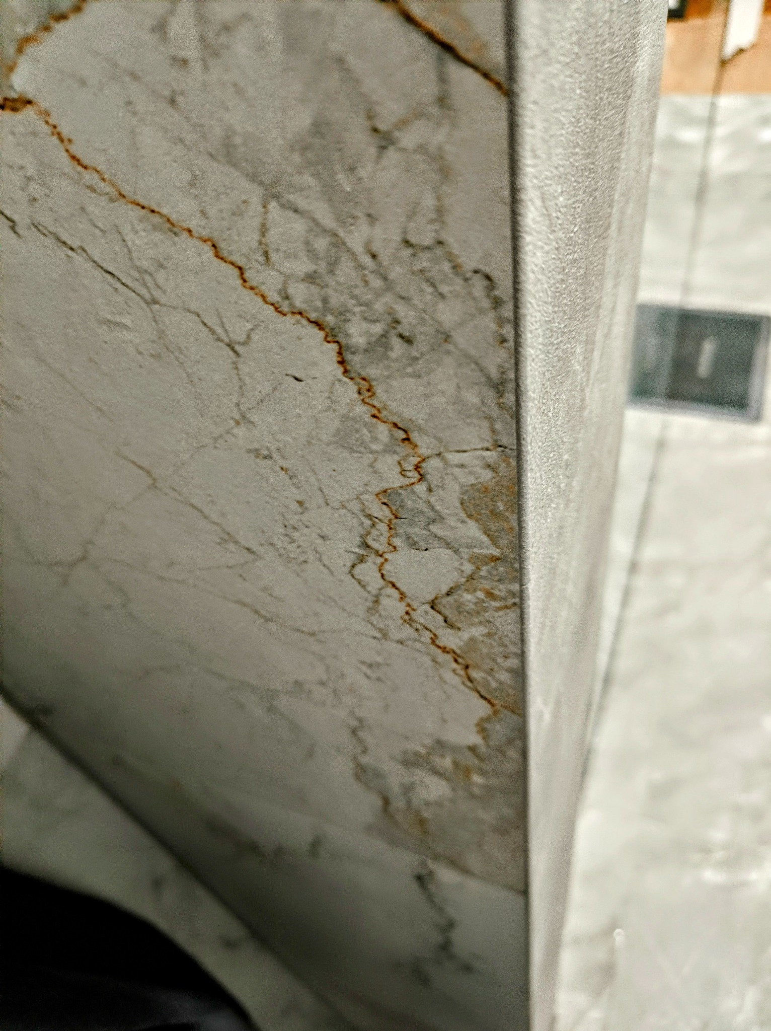 Fragment narożnika ściany wyłożonej płytkami imitującymi marmur z wyraźnymi, brązowymi żyłkami, widoczne fugi i fragment podłogi w tle.