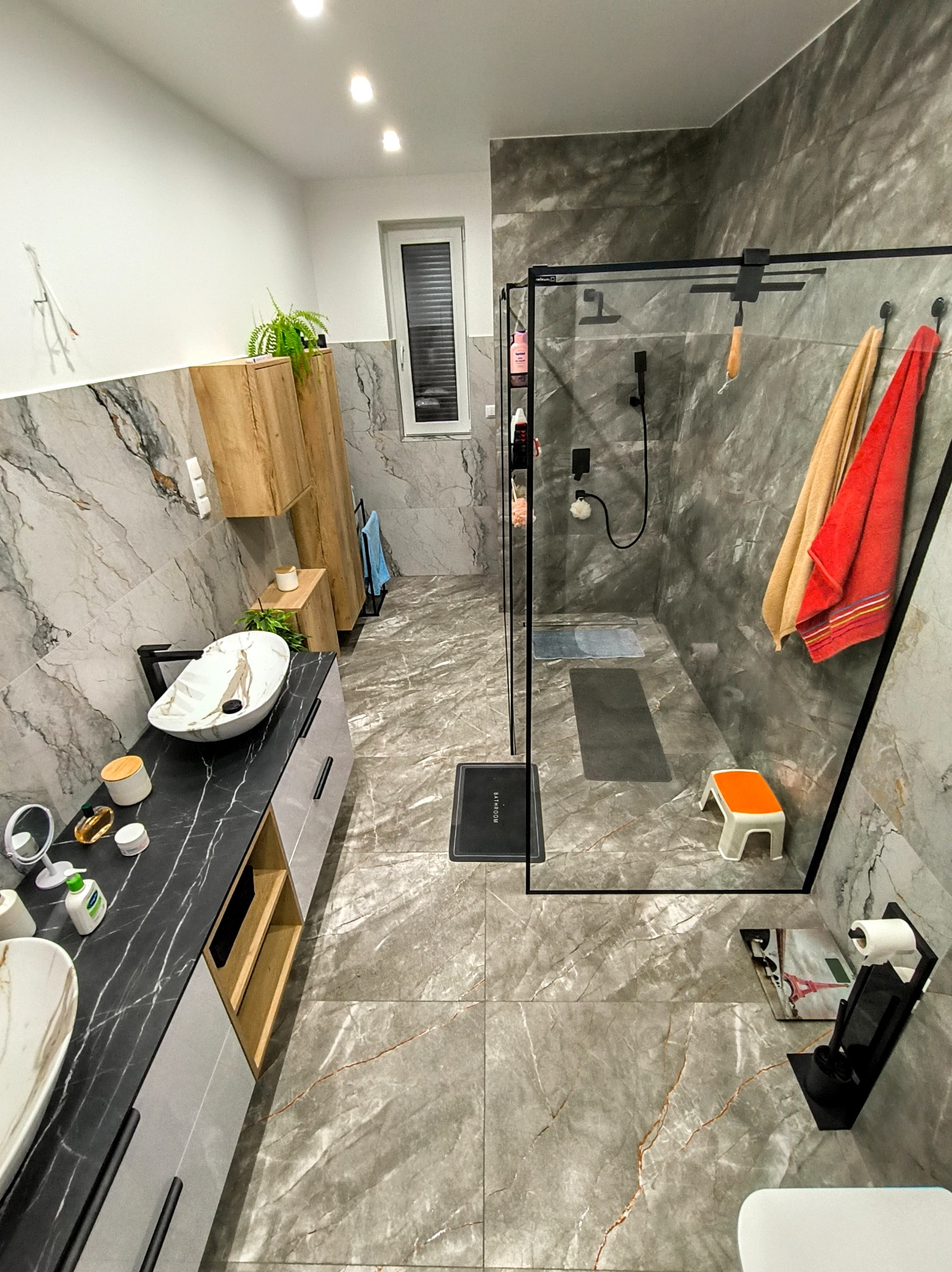 Nowoczesna łazienka z marmurowymi płytkami, kabiną prysznicową typu walk-in z czarną armaturą, umywalkami nablatowymi na czarnym blacie i drewnianymi szafkami, dopełniona pomarańczowym stołkiem...