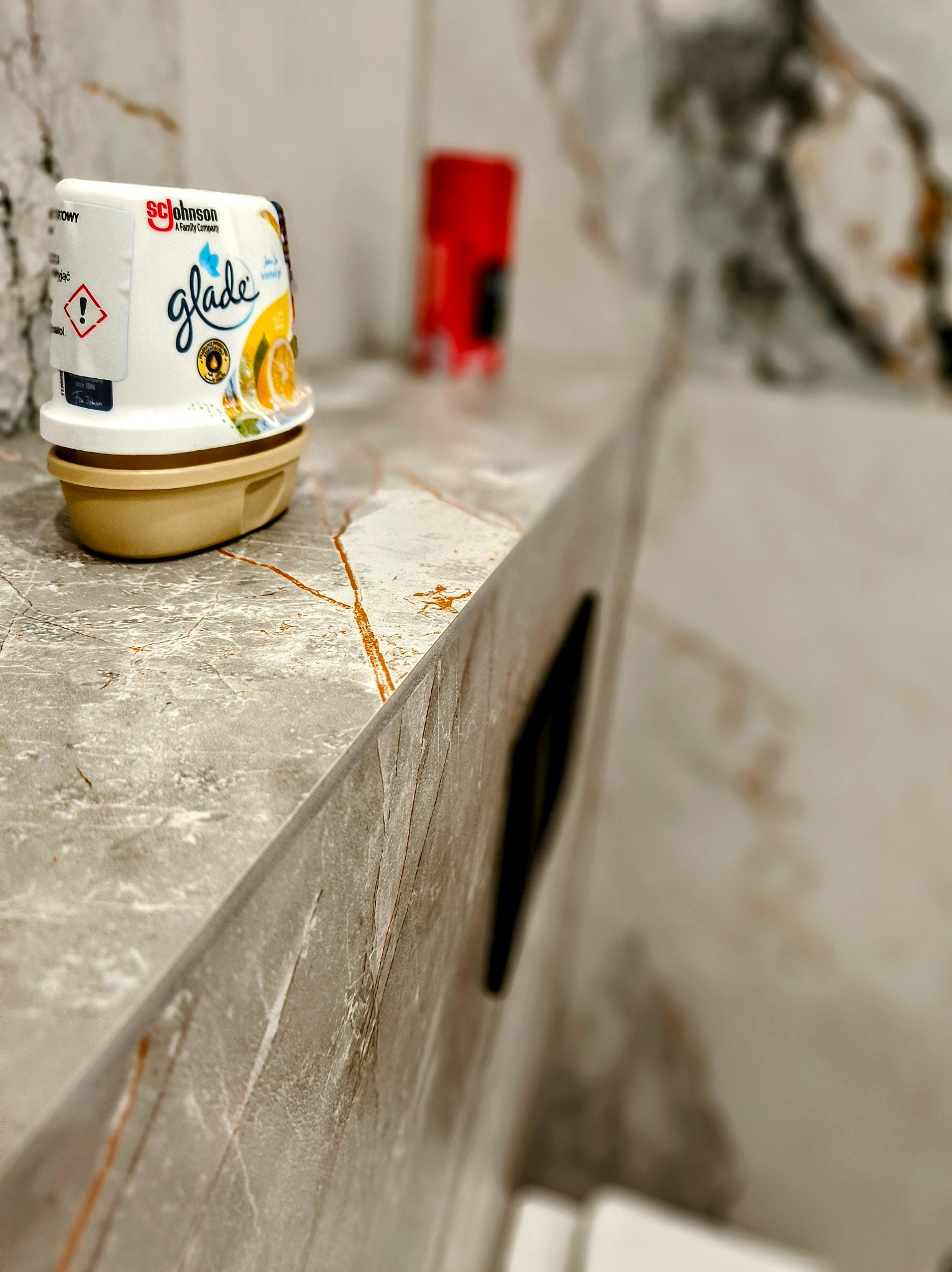 Półka z szarego marmuru z pomarańczowymi żyłkami w łazience w Gorzowie Wielkopolskim, na której stoi odświeżacz powietrza Glade z motywem pomarańczy. W tle widoczny czerwony pojemnik.