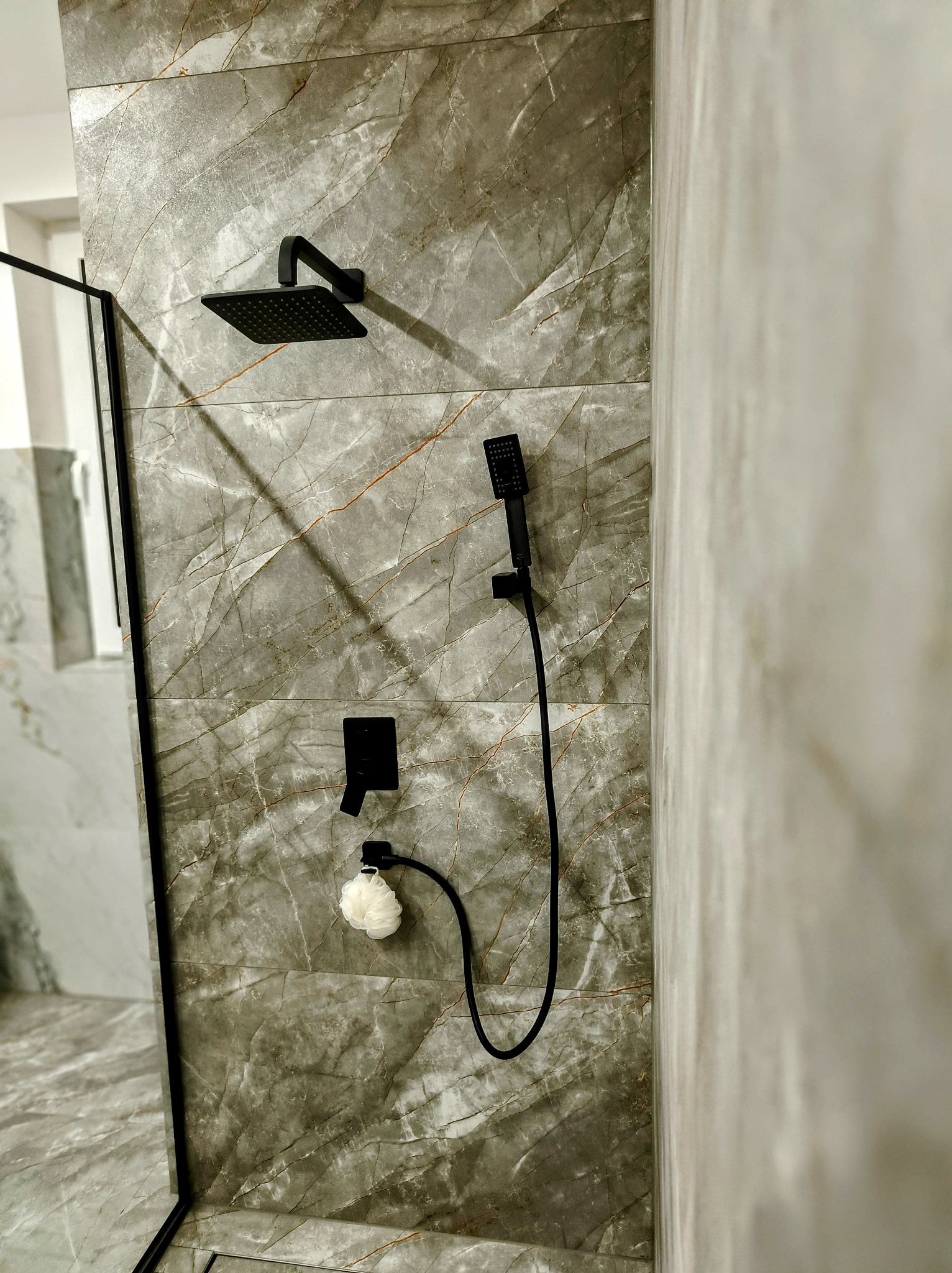 Szara, marmurkowa ściana kabiny prysznicowej z czarną armaturą: deszczownicą, słuchawką prysznicową i baterią, uzupełniona o białą myjkę.