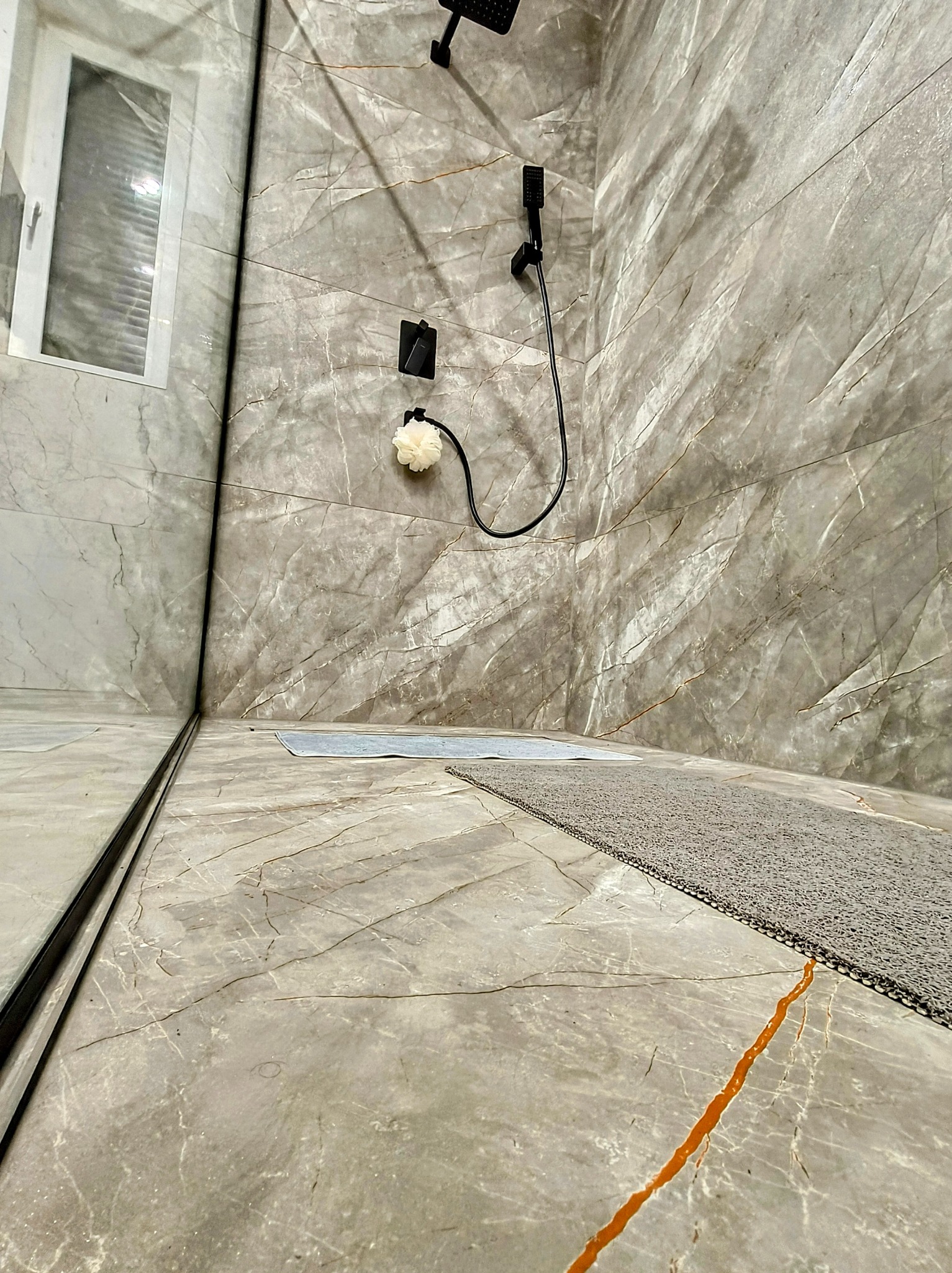 Kabina prysznicowa wykończona płytami imitującymi marmur, z czarną armaturą i beżową gąbką do mycia, widok z dołu.