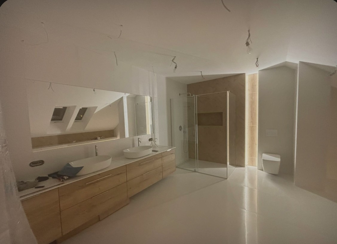 Nowoczesna łazienka w trakcie wykańczania, widoczne podwójne umywalki nablatowe na drewnianej szafce, przeszklona kabina prysznicowa i toaleta. Na suficie zwisające kable.