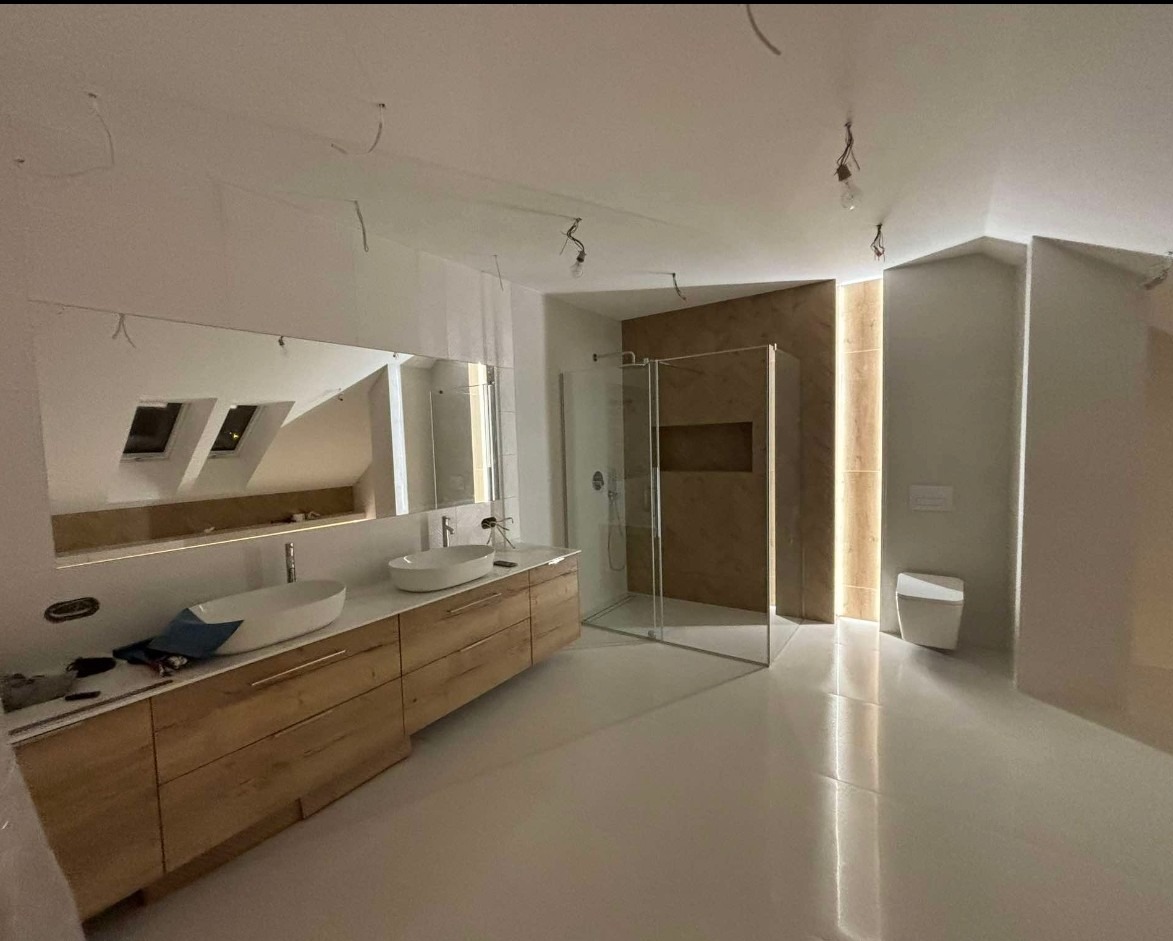 Wnętrze łazienki w trakcie wykańczania, widoczne podwieszane umywalki na drewnianej szafce, kabina prysznicowa z brązowymi płytkami i oświetleniem LED oraz wisząca toaleta, z widocznymi przewodami...