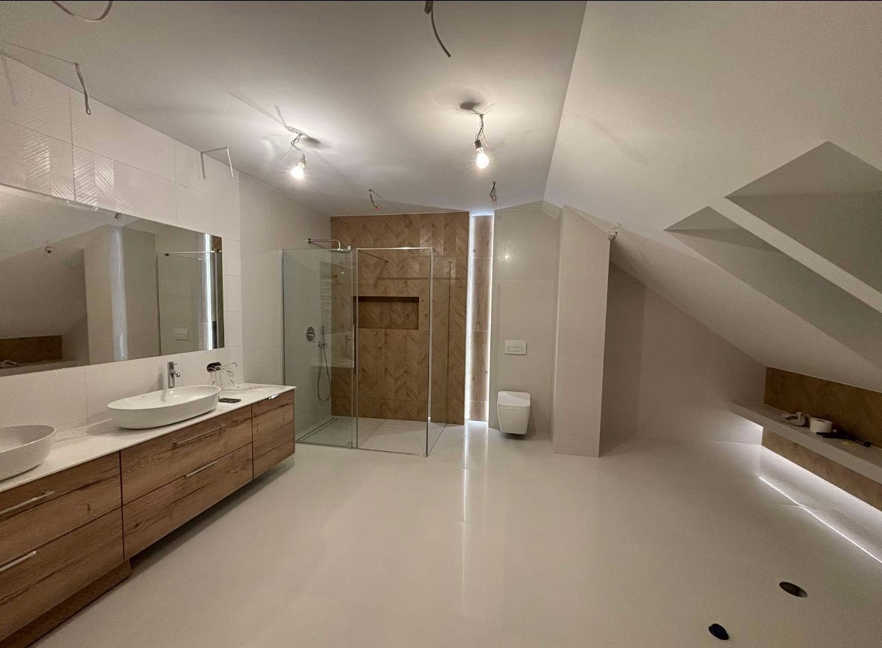 Przestronna łazienka na poddaszu z drewnianą szafką pod umywalką, szklaną kabiną prysznicową i akcentem drewnianej mozaiki na ścianie w strefie prysznica, oraz z widocznymi niedokończonymi pracami...