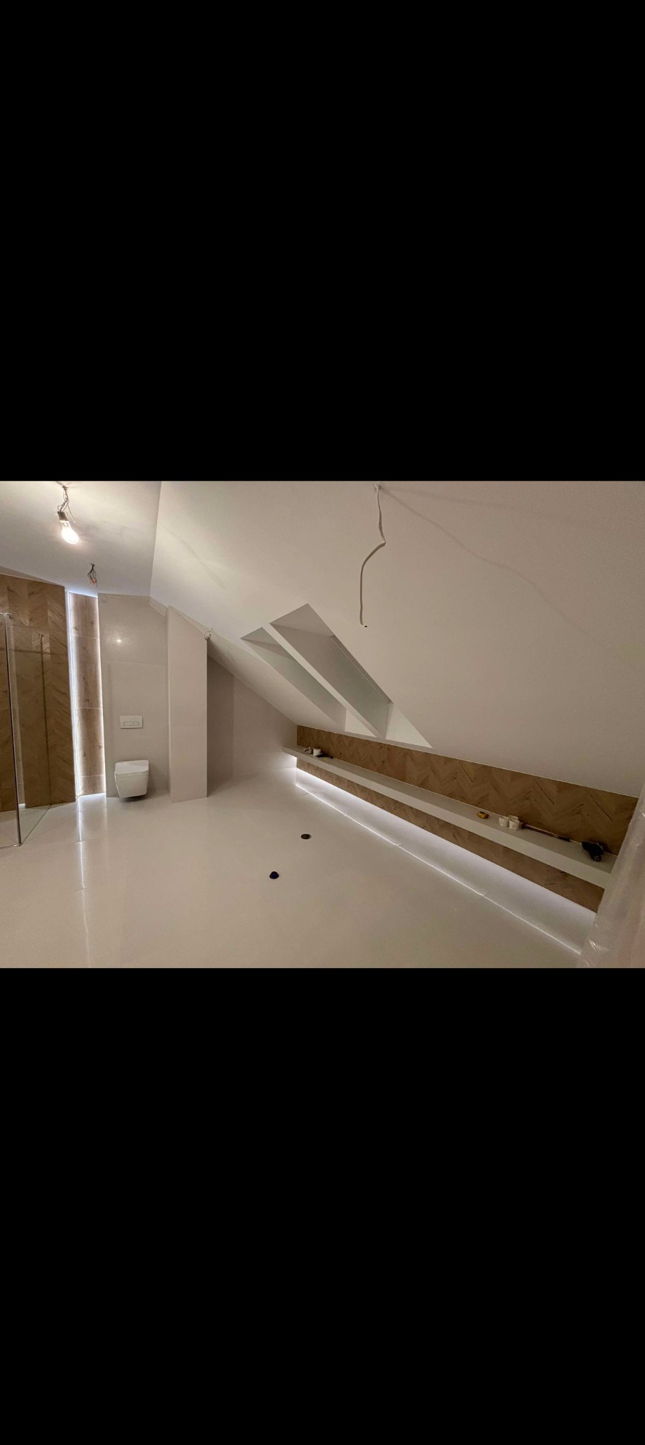 Wykończone poddasze ze skośnym sufitem, jasną podłogą i oświetloną półką wnękową, widoczna kabina prysznicowa i wisząca żarówka bez klosza.