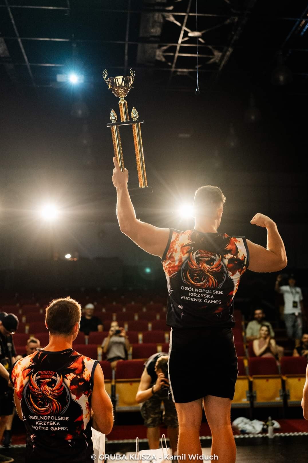 Mężczyzna z tyłu, w koszulce z logiem Phoenix Games, trzyma uniesiony puchar, prezentując muskulaturę na tle widowni. Scena z zawodów.