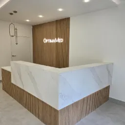Nowoczesna recepcja z marmurowym blatem i drewnianymi panelami ściennymi, podświetlonym logo 'OptimaMed' i minimalistyczną lampą wiszącą.