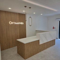 Kuźnia Mateusz Fiedorowicz - Nowoczesne wnętrze biura z recepcją wykończoną drewnianymi panelami i blatem z imitacji marmuru, oświetlone wiszącymi lampami i podświetlanym logo firmy na ścianie.