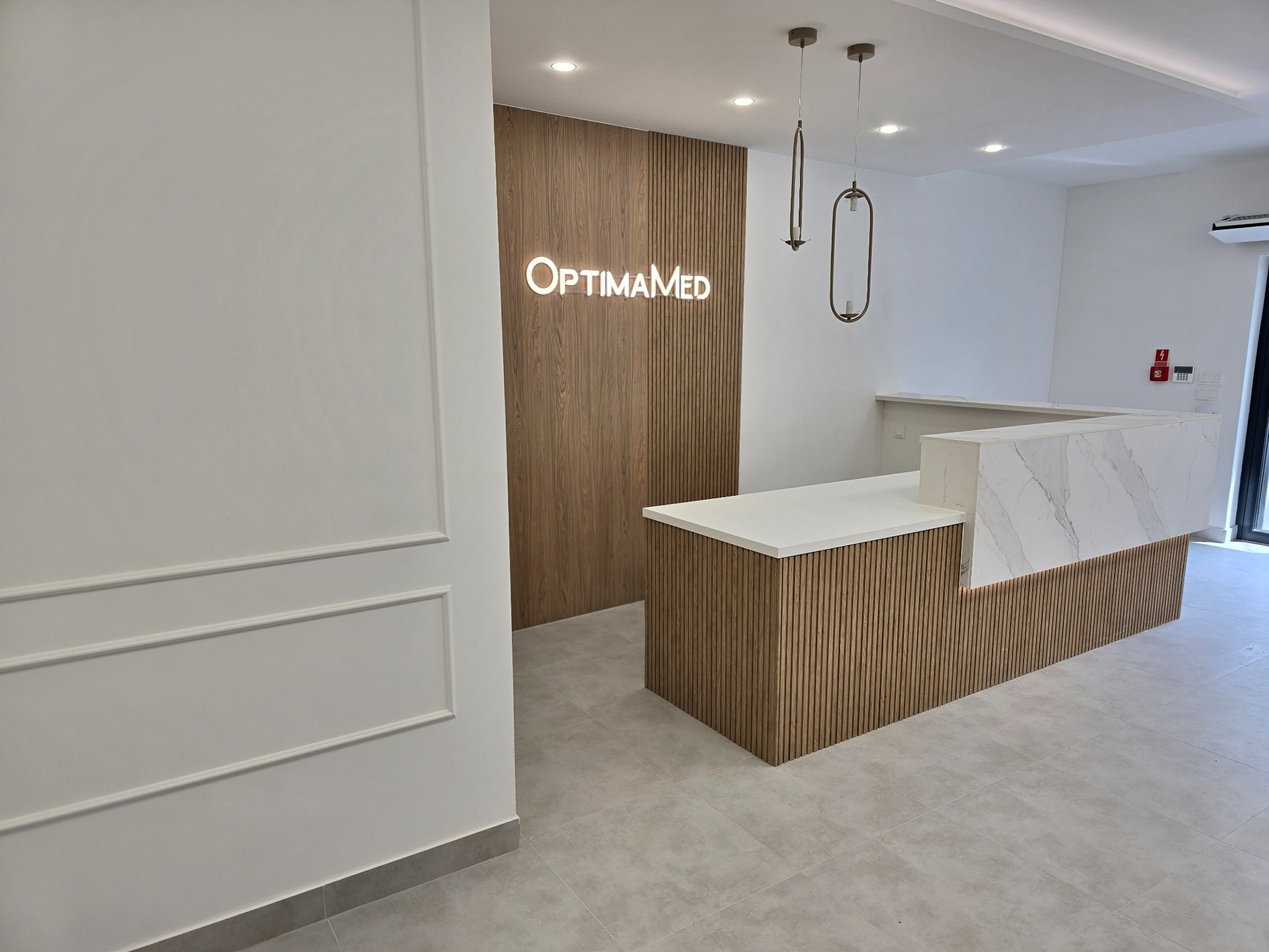 Nowoczesne wnętrze recepcji z drewnianą ścianą z podświetlonym logo OptimaMed, marmurowym blatem i minimalistycznymi lampami wiszącymi.