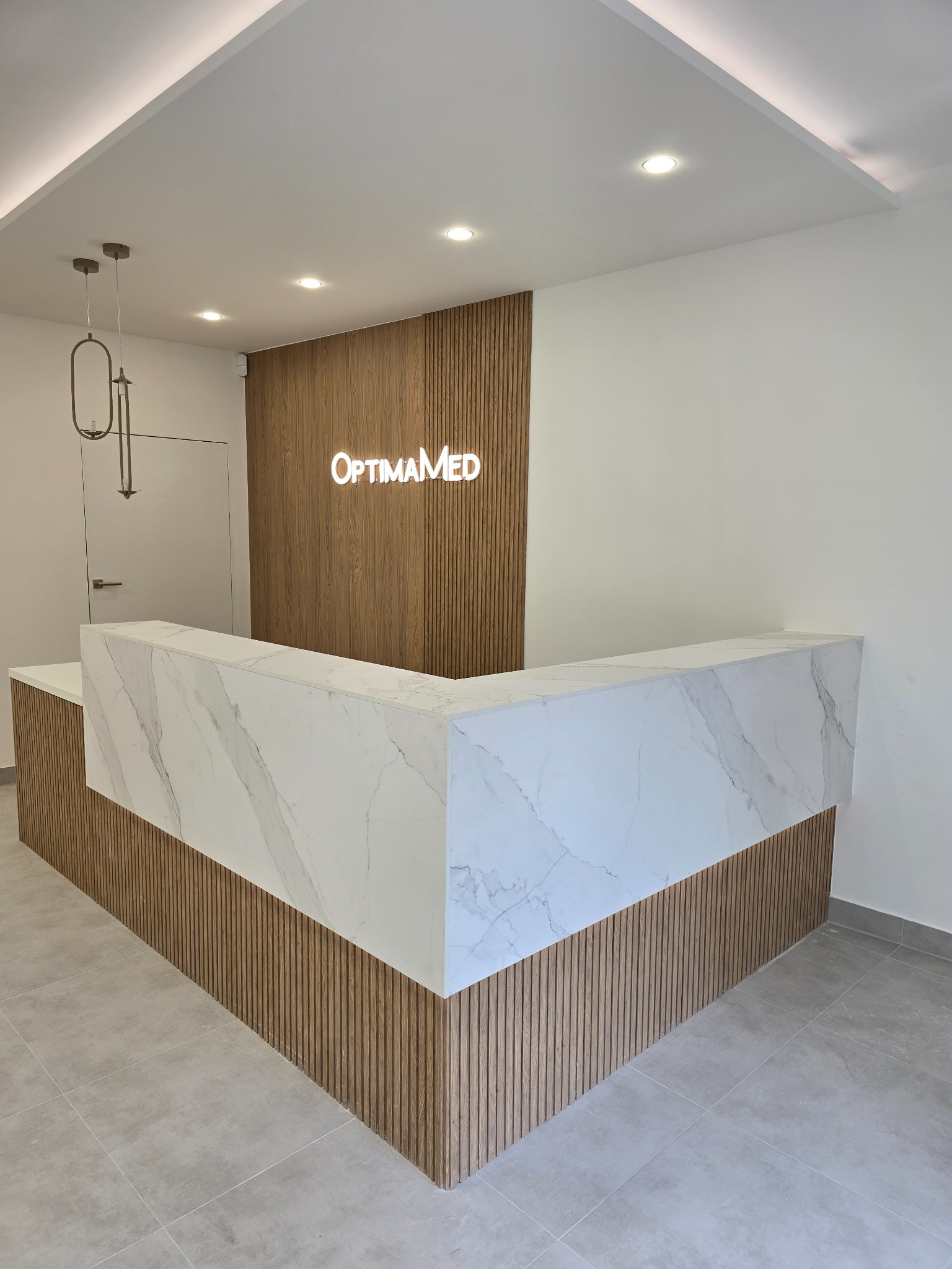 Nowoczesna recepcja z marmurowym blatem i drewnianymi panelami ściennymi, podświetlonym logo 'OptimaMed' i minimalistyczną lampą wiszącą.