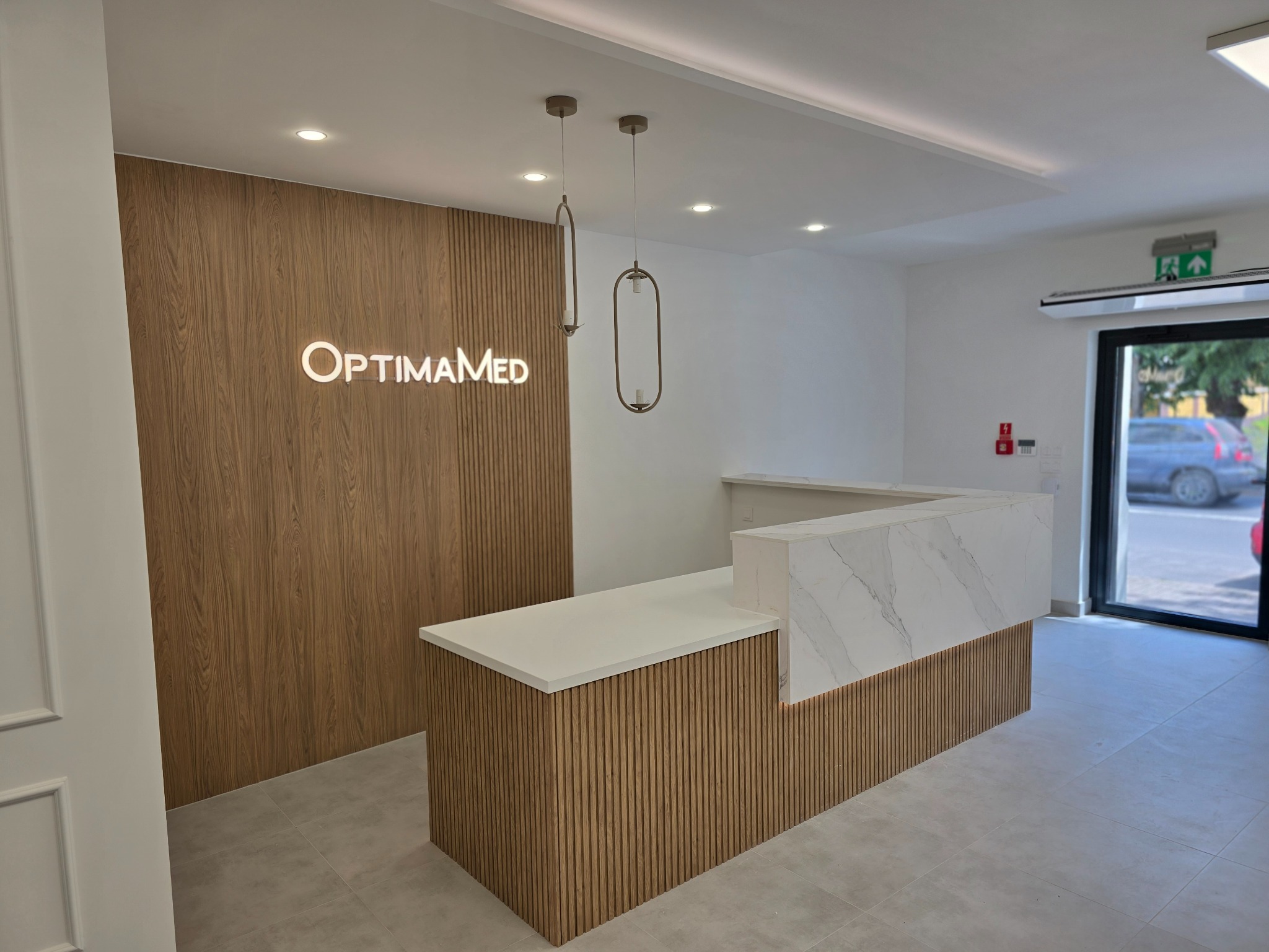 Nowoczesne wnętrze biura z recepcją wykończoną drewnianymi panelami i blatem z imitacji marmuru, oświetlone wiszącymi lampami i podświetlanym logo firmy na ścianie.