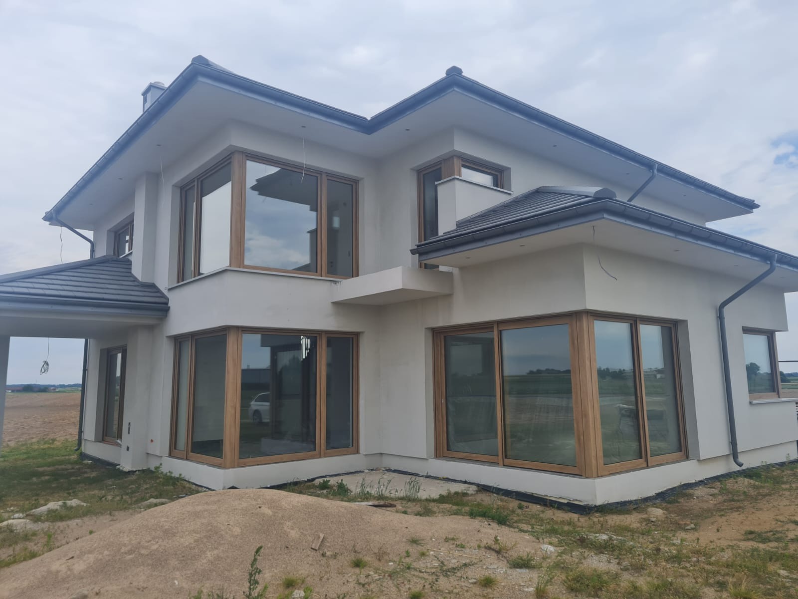 Nowoczesny dom murowany z dużymi oknami w drewnianych ramach, szary dach, jeszcze niezagospodarowany teren wokół.