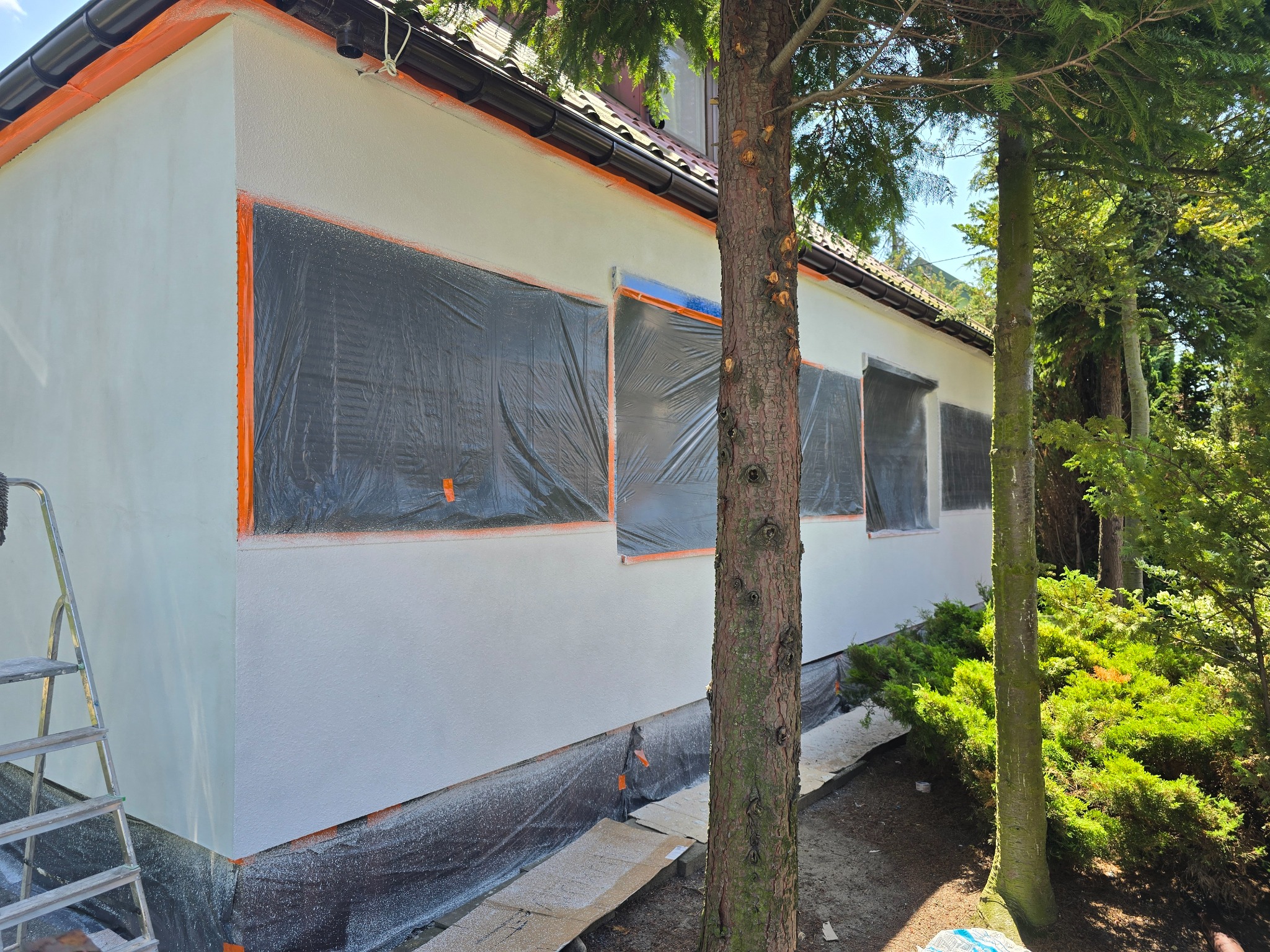 Ocieplanie elewacji domu jednorodzinnego z zabezpieczonymi oknami folią i taśmą malarską, widoczna drabina i drzewo w pierwszym planie.
