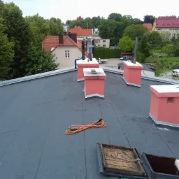 Widok z góry na płaski dach pokryty papą, z widocznymi kominami pomalowanymi na różowo, zwiniętym pomarańczowym przedłużaczem, otwartym włazem dachowym z drewnianą ramą i panoramą miasta w tle.