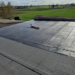 Płaski dach pokryty papą termozgrzewalną z widocznym narzędziem do zgrzewania i zielonym krajobrazem w tle.