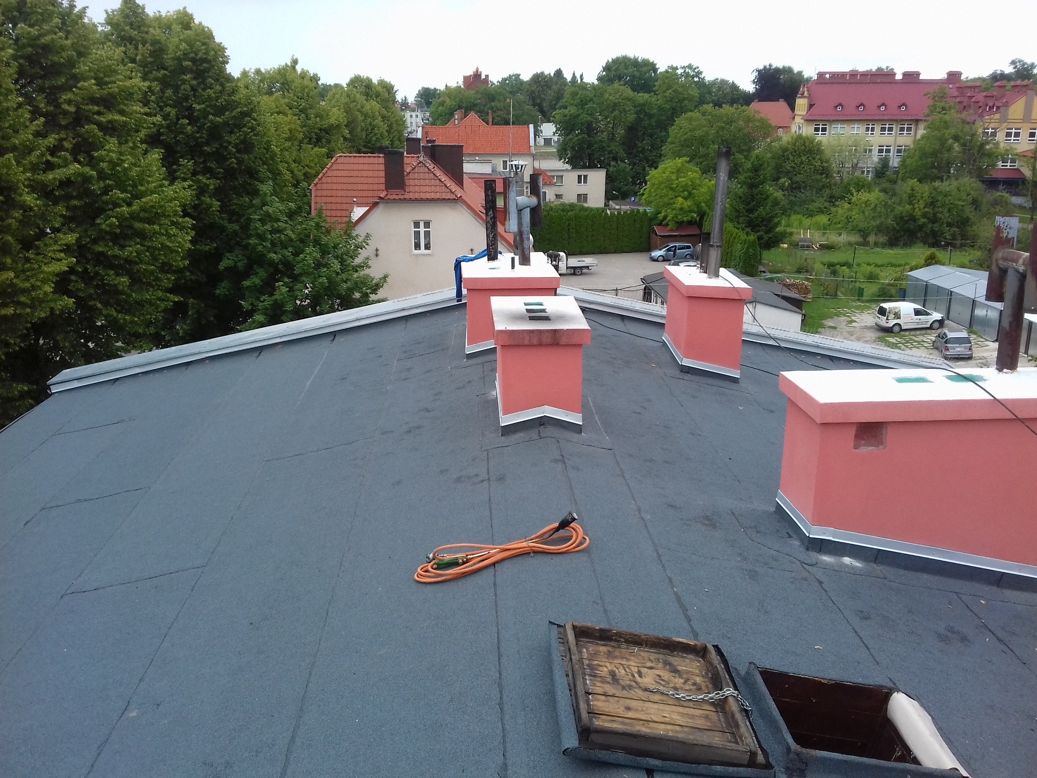 Widok z góry na płaski dach pokryty papą, z widocznymi kominami pomalowanymi na różowo, zwiniętym pomarańczowym przedłużaczem, otwartym włazem dachowym z drewnianą ramą i panoramą miasta w tle.