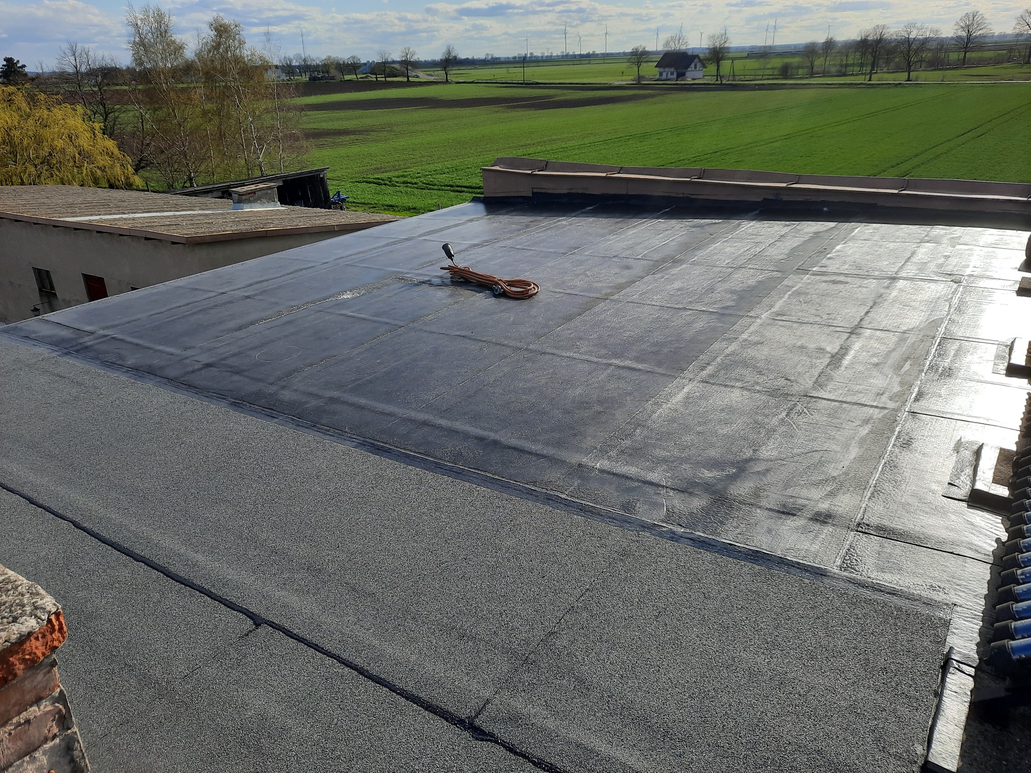 Płaski dach pokryty papą termozgrzewalną z widocznym narzędziem do zgrzewania i zielonym krajobrazem w tle.