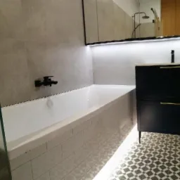 Nowoczesna łazienka z białą wanną obudowaną płytkami, czarną armaturą, szafką pod umywalką i podświetlanym lustrem, wzorzysta podłoga.