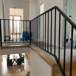 Nowoczesna czarna balustrada metalowa z drewnianym wykończeniem na antresoli, widok na jadalnię z drewnianą podłogą, w tle pomieszczenie w trakcie wykańczania.