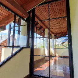 Przeszklona weranda z czarnymi, metalowymi ramami okiennymi, widok z zewnątrz na brązową podłogę z płytek i drewniany sufit, kolumny w tle.