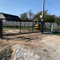 Czarna metalowa brama przesuwna z siatką, częściowo zamontowana, z małą żółtą koparką w tle i betonowym fundamentem w surowym otoczeniu.