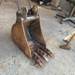 Używana, zardzewiała łyżka koparki z trzema zębami, leżąca na betonowej posadzce warsztatu, widoczny młotek i wózek narzędziowy w tle.