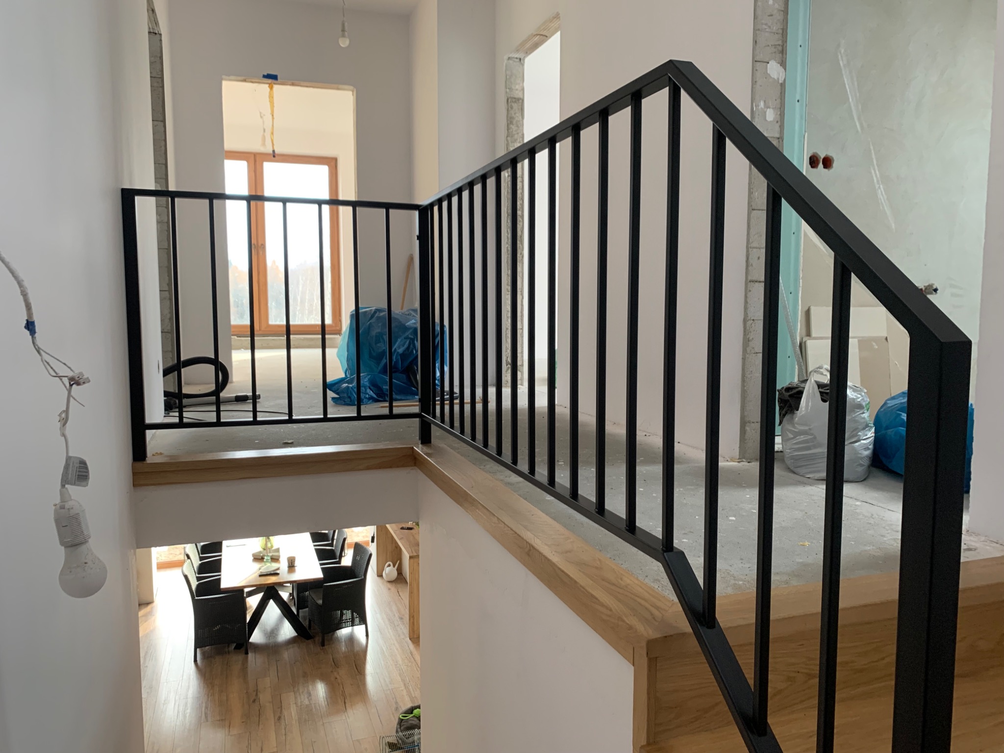 Nowoczesna czarna balustrada metalowa z drewnianym wykończeniem na antresoli, widok na jadalnię z drewnianą podłogą, w tle pomieszczenie w trakcie wykańczania.