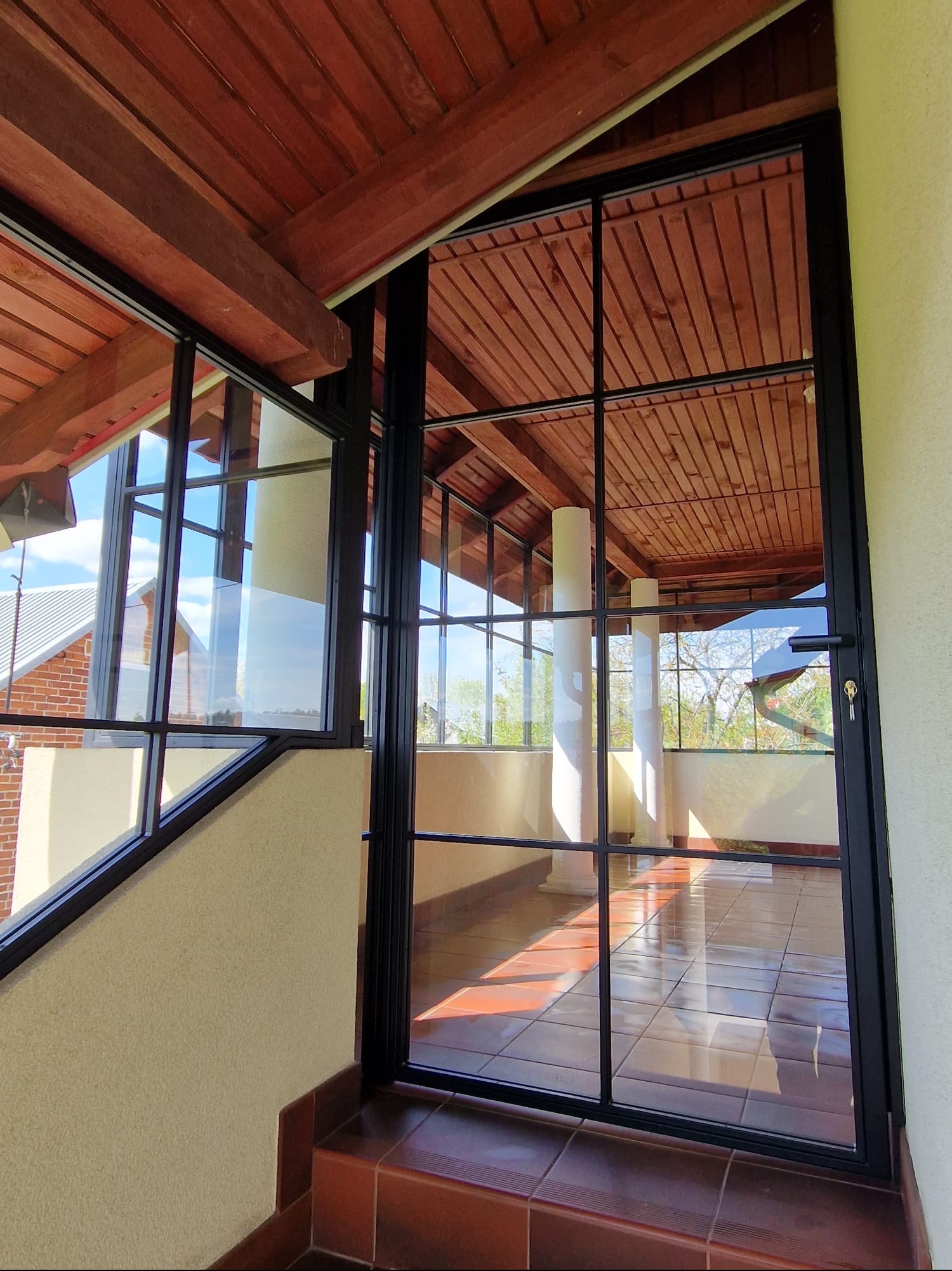 Przeszklona weranda z czarnymi, metalowymi ramami okiennymi, widok z zewnątrz na brązową podłogę z płytek i drewniany sufit, kolumny w tle.