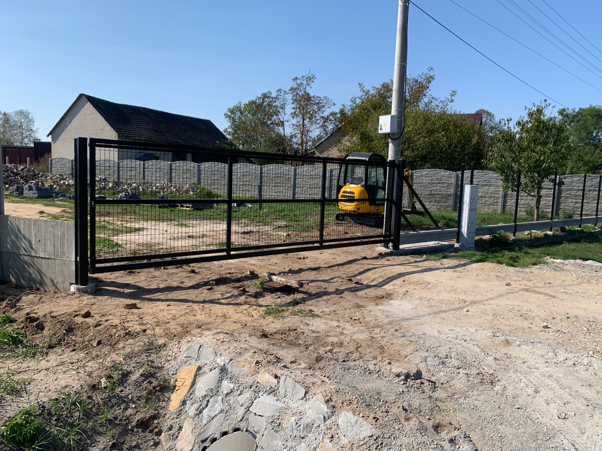 Czarna metalowa brama przesuwna z siatką, częściowo zamontowana, z małą żółtą koparką w tle i betonowym fundamentem w surowym otoczeniu.