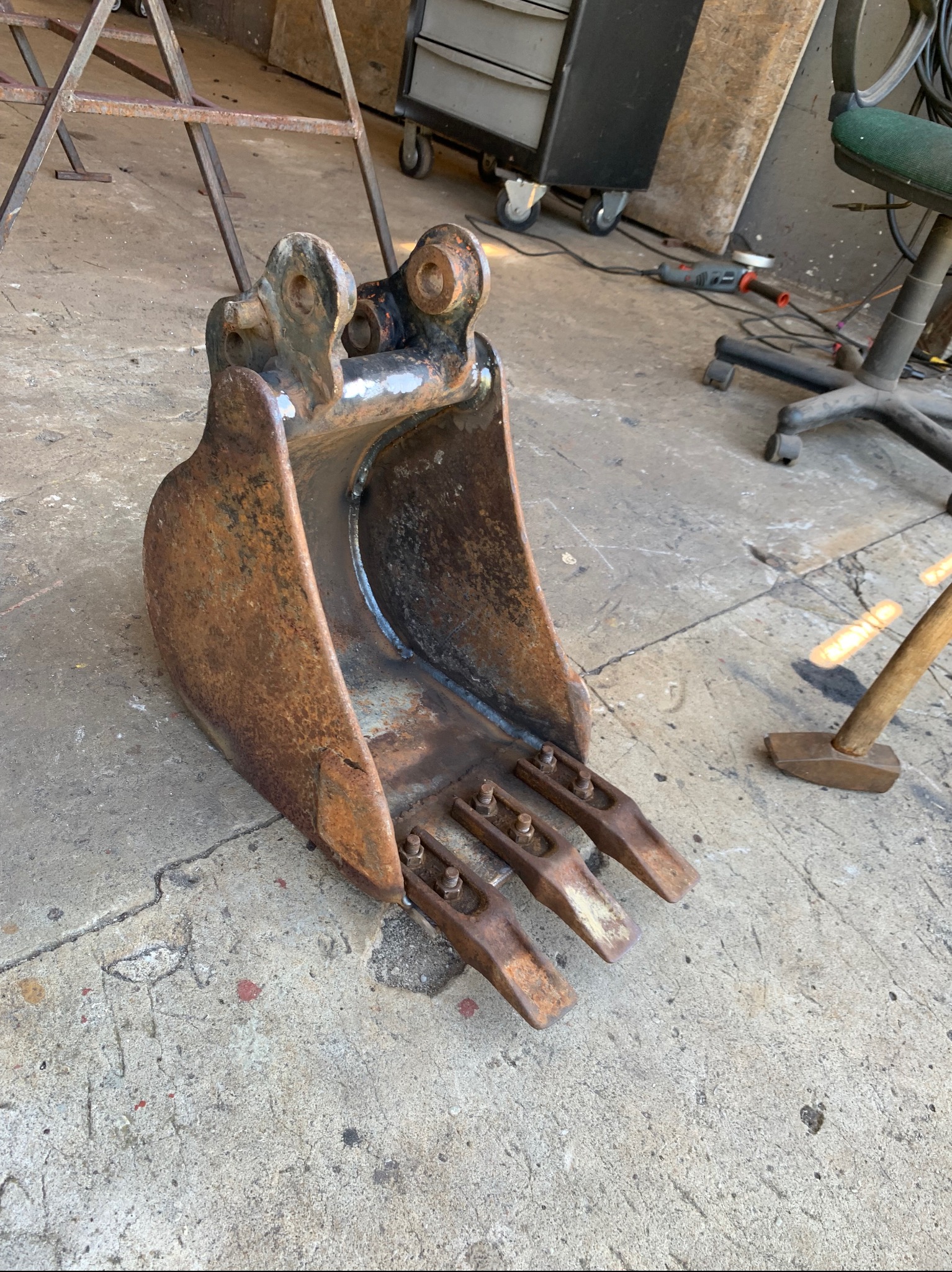 Używana, zardzewiała łyżka koparki z trzema zębami, leżąca na betonowej posadzce warsztatu, widoczny młotek i wózek narzędziowy w tle.