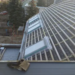 Konstrukcja dachu z zamontowanymi oknami dachowymi w trakcie budowy, widoczne łaty, membrana dachowa, narzędzia dekarskie i otoczenie domu.