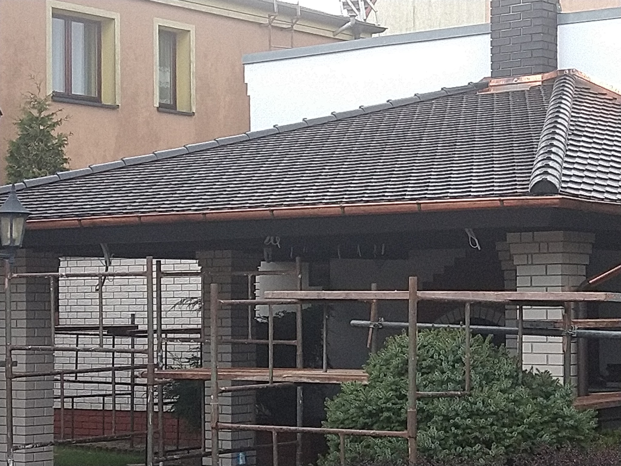 Nowy dach z ciemnej dachówki ceramicznej, widoczne miedziane rynny i obróbki blacharskie przy kominie z cegły klinkierowej, rusztowanie w tle.