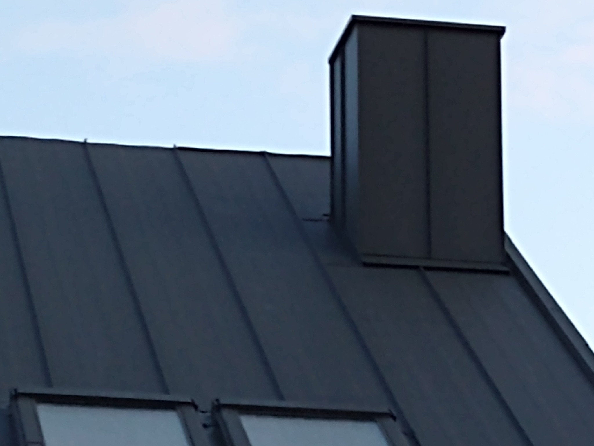 Ciemny, metalowy dach z oknami dachowymi i kominem w kształcie prostopadłościanu, widoczny na tle jasnego nieba.