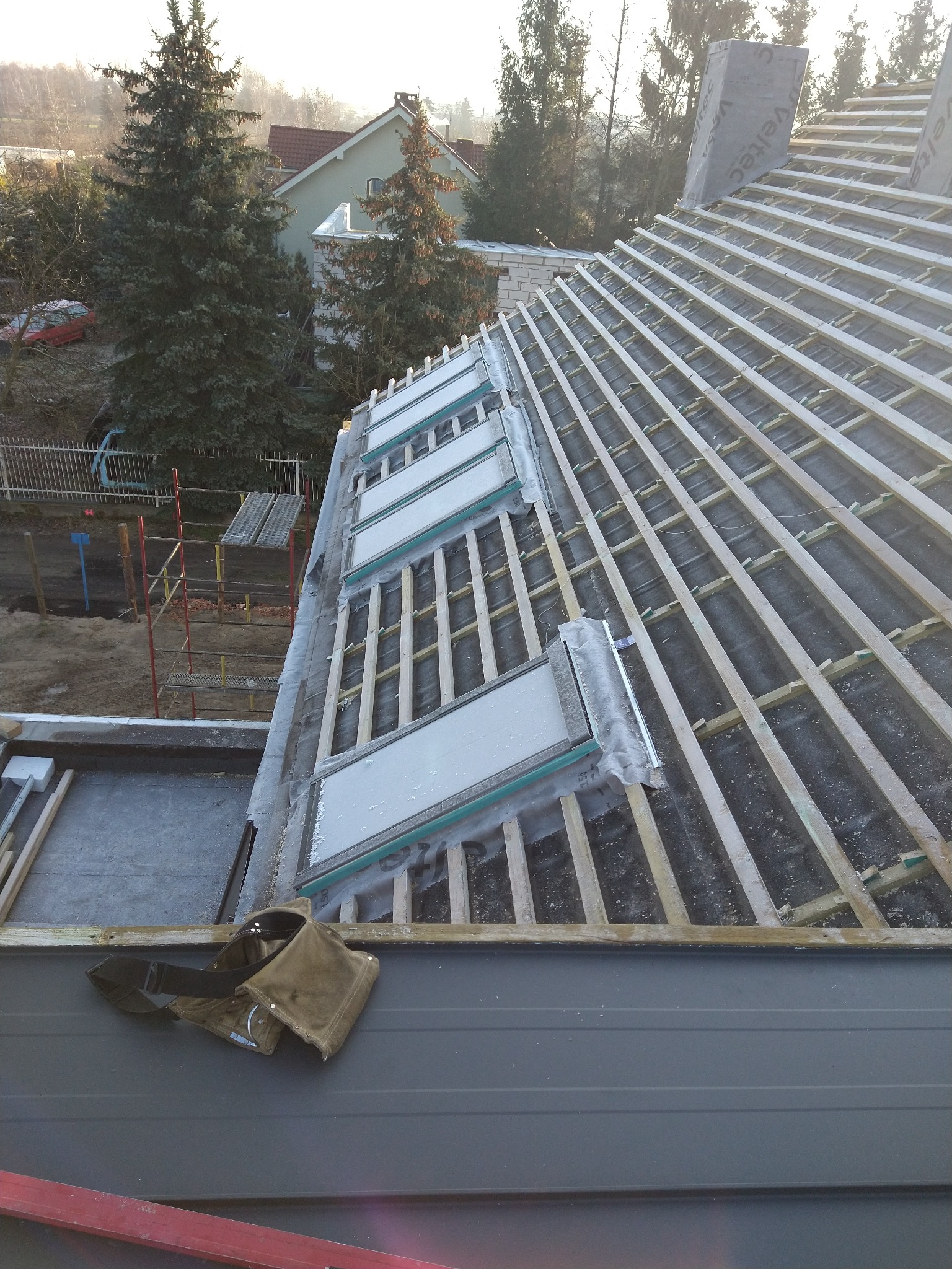 Konstrukcja dachu z zamontowanymi oknami dachowymi w trakcie budowy, widoczne łaty, membrana dachowa, narzędzia dekarskie i otoczenie domu.