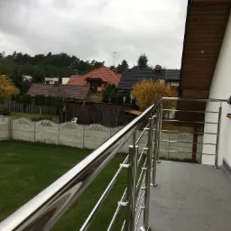 Balkon z polerowaną, metalową balustradą o minimalistycznym designie, z widokiem na trawnik, ogrodzenie z prefabrykatów betonowych i zabudowę jednorodzinną w tle, pochmurne niebo.
